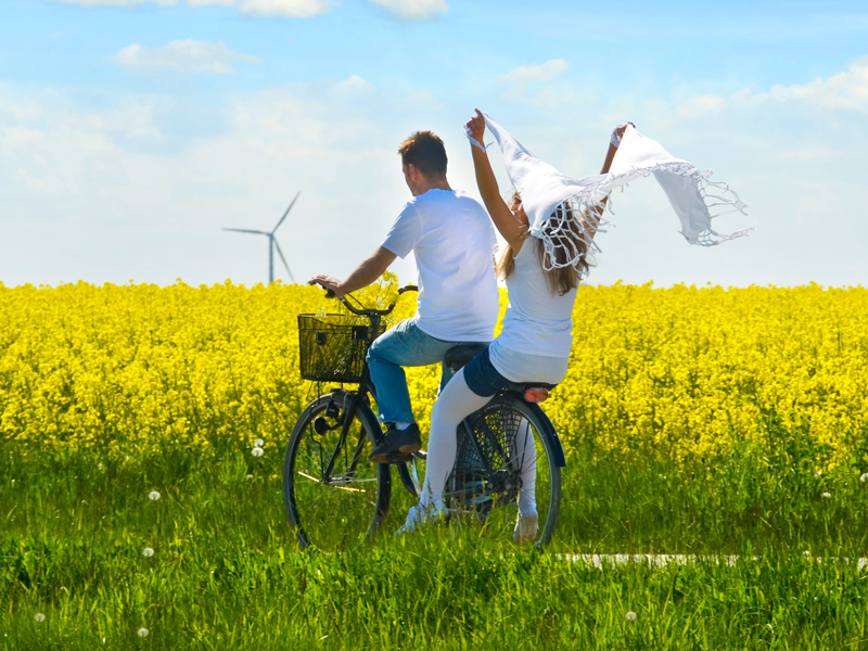 to unge mennesker på en cykel gennem det danske sommerlandskab med en rapsmark og en vindmølle i baggrunden.