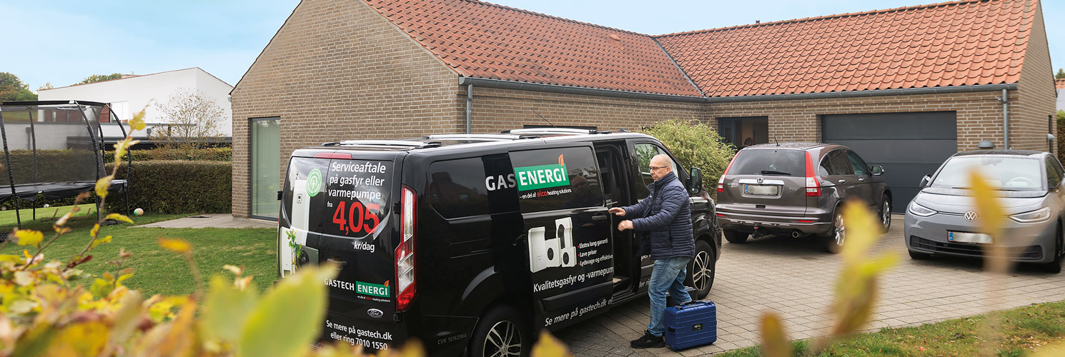Servicemontør fra gastech-energi står i en indkørsel til t parcelhus og er ved at tage ting ud af sin bil, så han kan gå ind i huset og servicere et gasfyr eller en varmepumpe.
